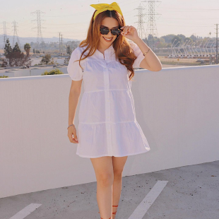 Stylish young woman is shown outside on a parking garage deck wearing a white babydoll dress and sunglasses