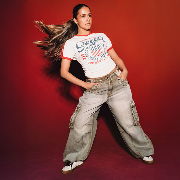 Person in a white USA soccer graphic tee and cargo pants posing in motion against a red background.