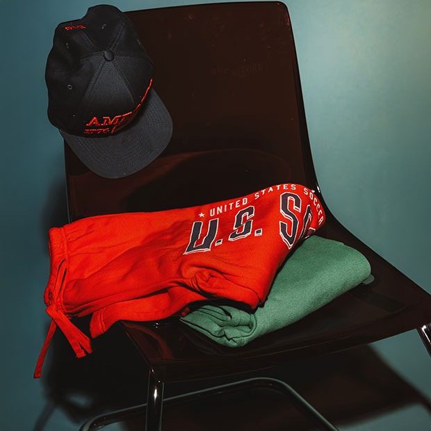 Red USA sweatpants, green sweatshirt, and black cap arranged on a chair against a green backdrop.