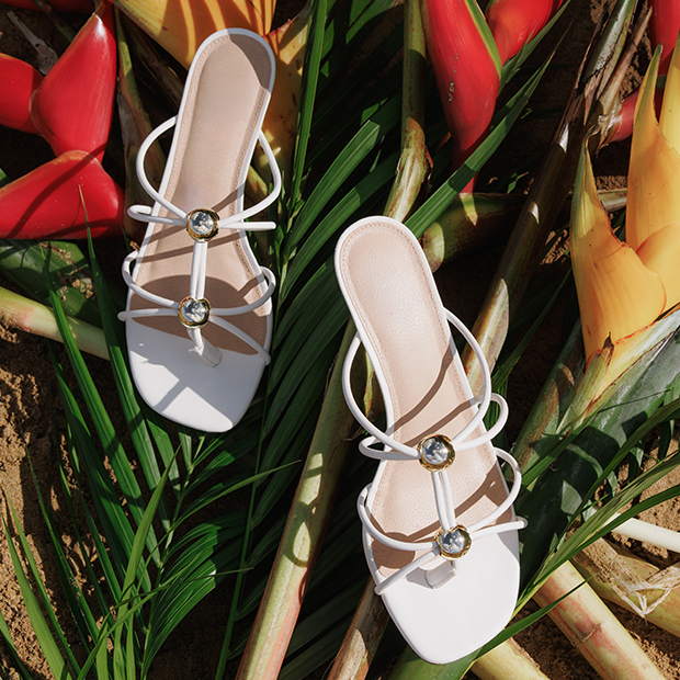 White strappy sandals with jeweled details placed among tropical flowers and greenery.