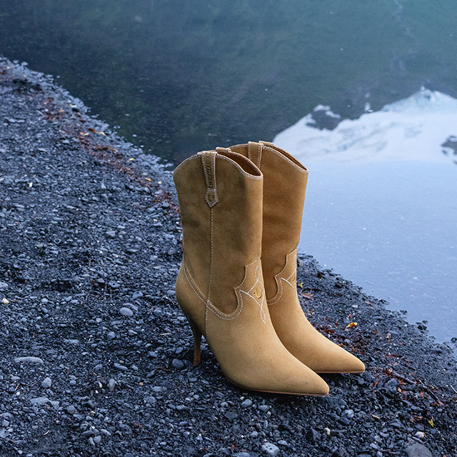 Pair of tan suede western ankle boots with pointed toes and high heels displayed on a rocky lakeshore.